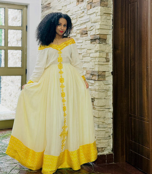 A woman wearing a long white and yellow embroidered Habesha Dress stands indoors next to a stone wall and dark wooden door, holding out the sides of the full skirt to show its shape.