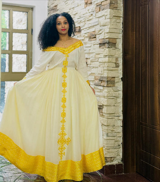 A woman wearing a long cream and yellow embroidered Habesha Dress, holding out the skirt to show its full volume, standing in front of a stone wall and dark wooden door.