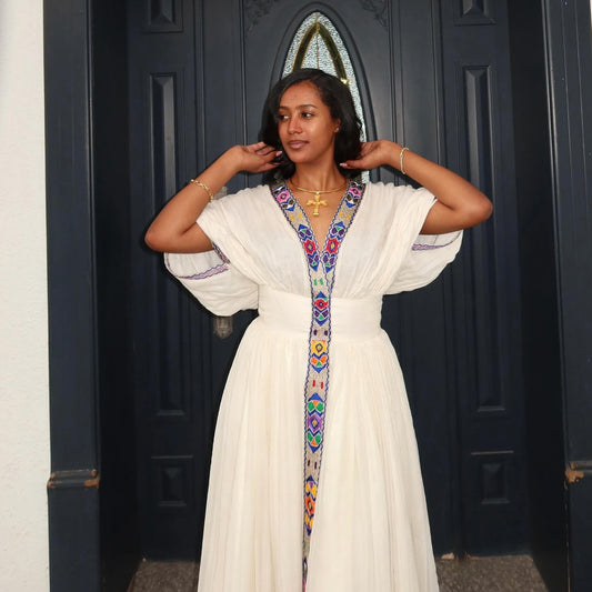 A woman wearing a white long Habesha Kemis  with colorful embroidered trim stands in front of a dark blue door.