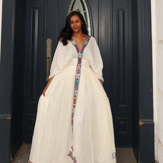 Woman in a white Habesha Kemis  with colorful embroidery standing in front of a dark blue door.