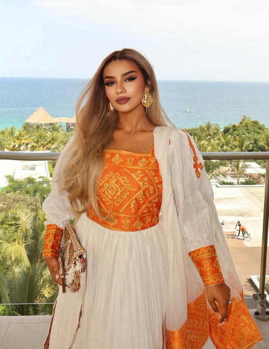 Woman in an orange and white traditional habesha kemis standing on a balcony with ocean view
