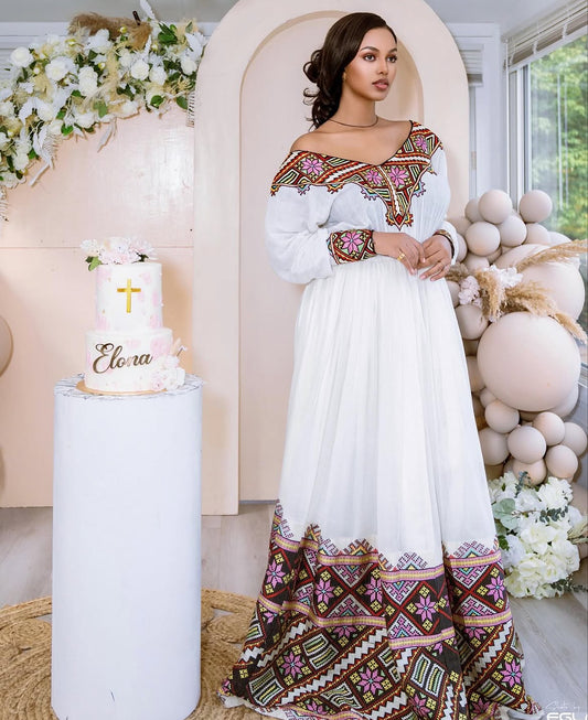 Woman in a white and patterned dress standing next to a cake with 'Eloisa' on it, surrounded by decorative elements.