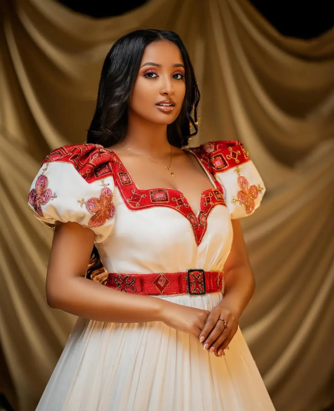 Woman wearing a Habesha Wedding Dress with red and white embroidery against a beige curtain background