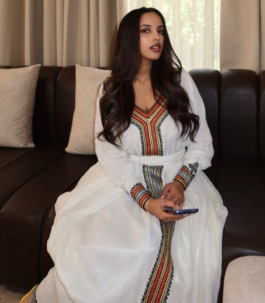 A woman wearing a white traditional Habesha Kemis with colorful embroidered patterns sits on a dark brown leather sofa.
