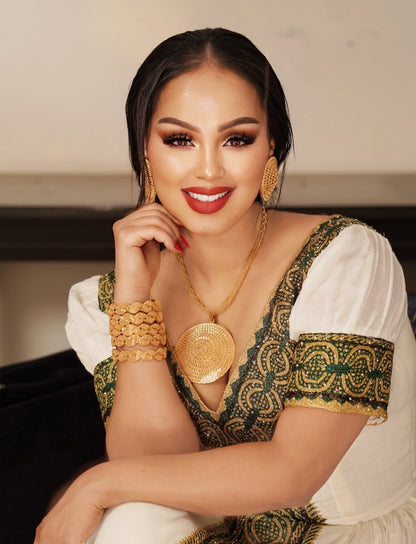 Woman wearing Habesha Dress attire with gold jewelry against a neutral background