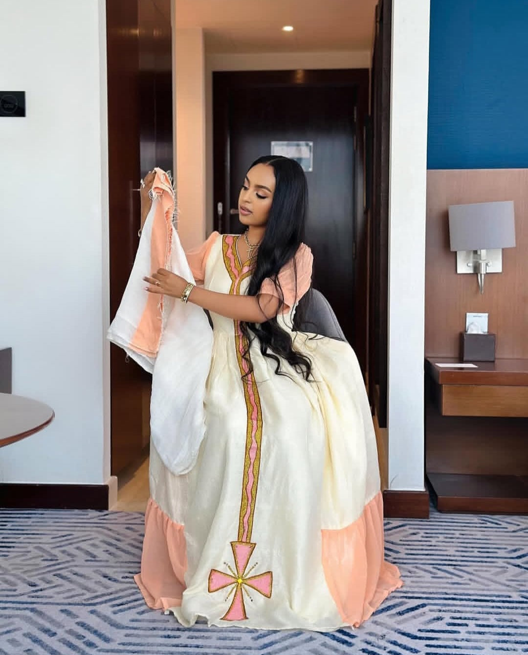 Woman holding a traditional Ethiopian Dress in a room with a blue wall and wooden furniture.