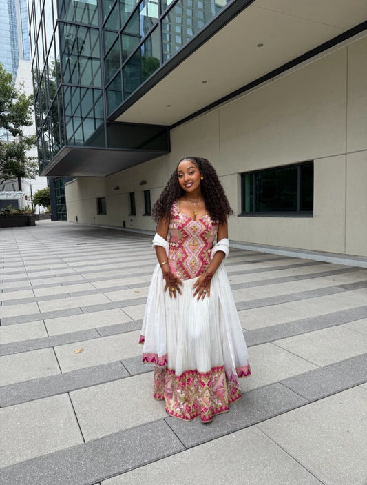 A woman stands outdoors in front of a modern glass building, wearing a long white Habesha Dress with pink and gold geometric embroidery on the bodice and a pink patterned hem.