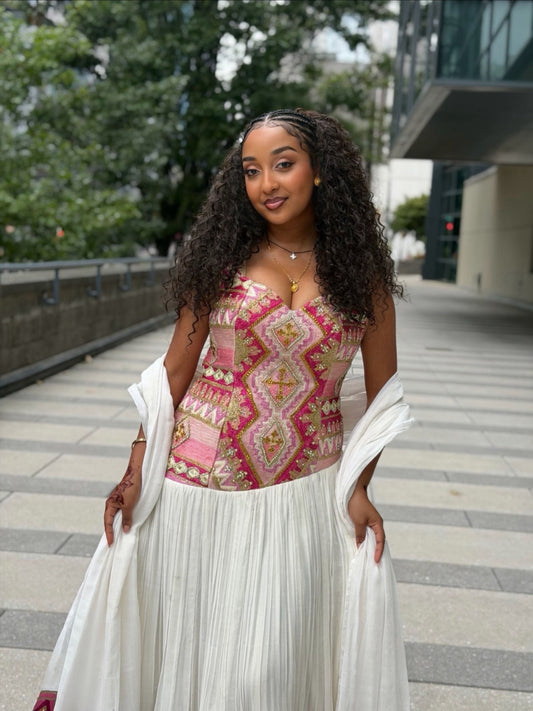 A woman wearing a pink and gold patterned corset-style Habesha Dress with a long white pleated skirt and draped white sleeves, standing outdoors on a paved walkway with trees and a modern building in the background.