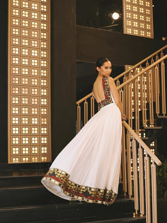 Woman in a white Habesha zuria with red and gold patterns standing on a staircase at night.
