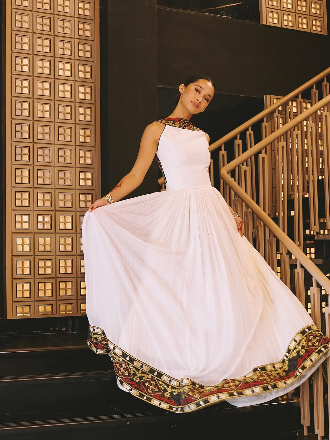 Woman in a white habesha dress with decorative hem standing on a staircase.