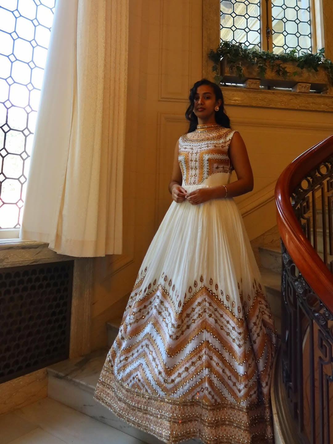 Woman in a white and gold patterned dress standing in a room with wooden paneling and a staircase.