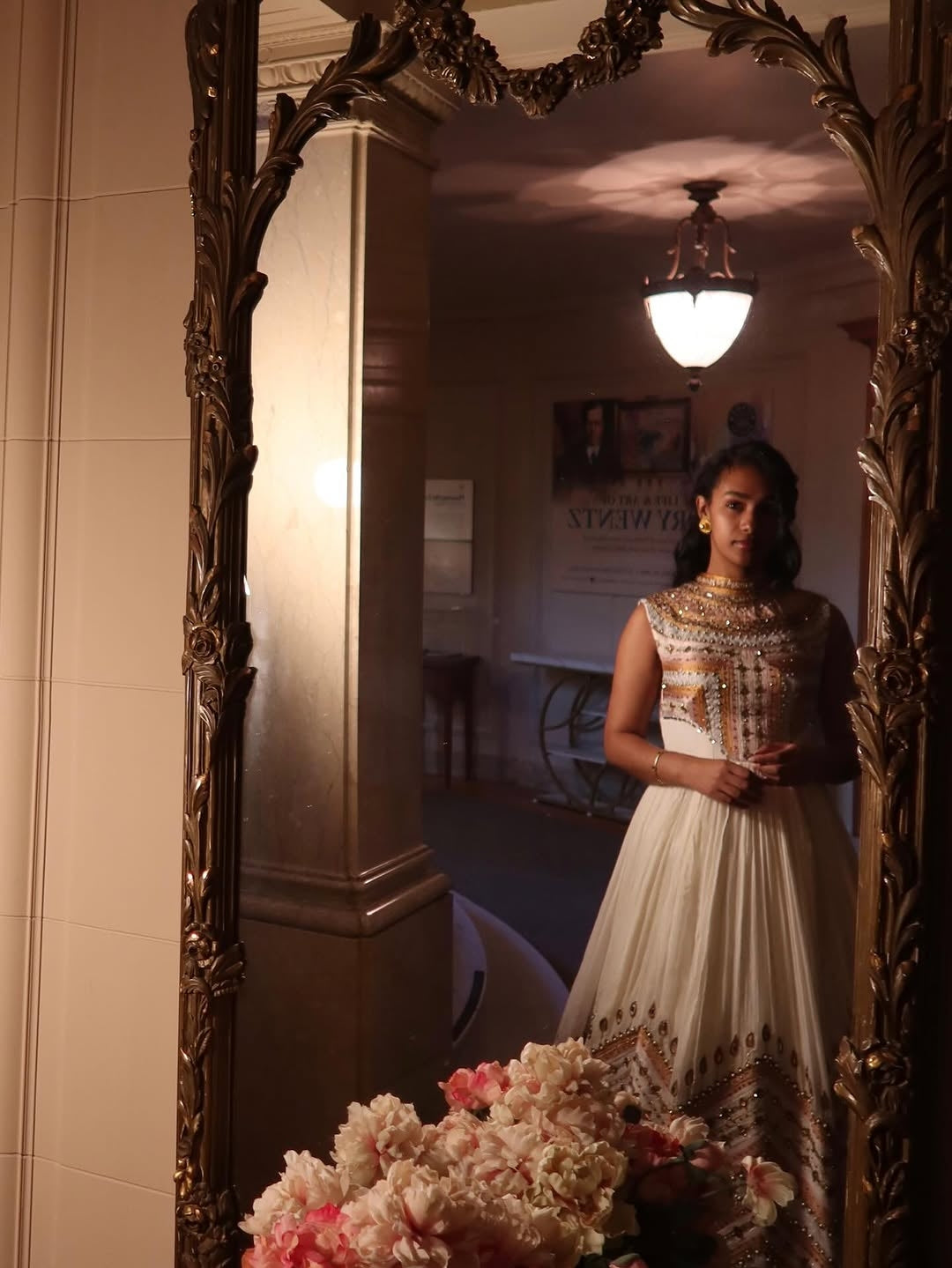 Woman in a white and gold dress standing in front of an ornate mirror with floral decorations.