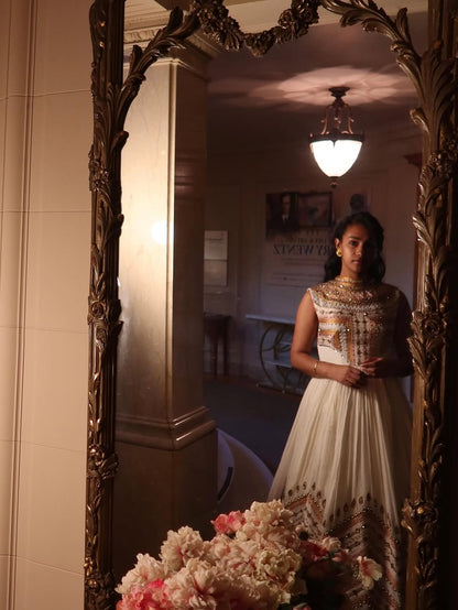 Woman in a white and gold dress standing in front of an ornate mirror with floral decorations.