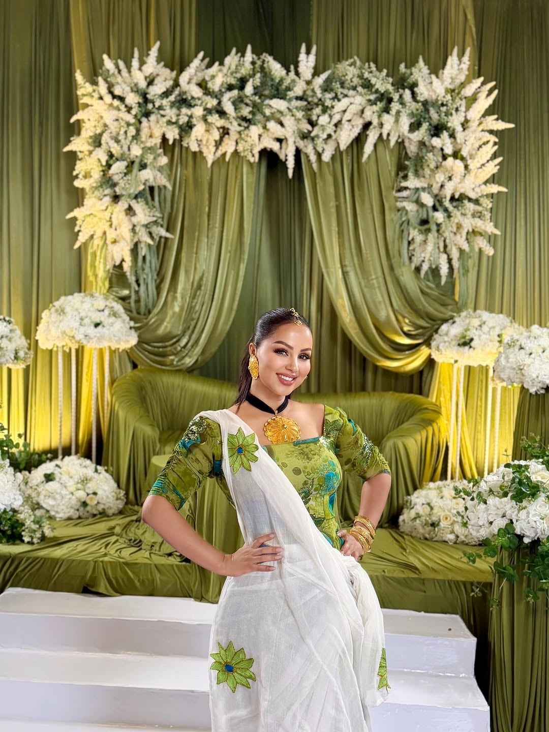 Woman in a green and white traditional Shiffon Habesha standing in front of floral decorations and green curtains.