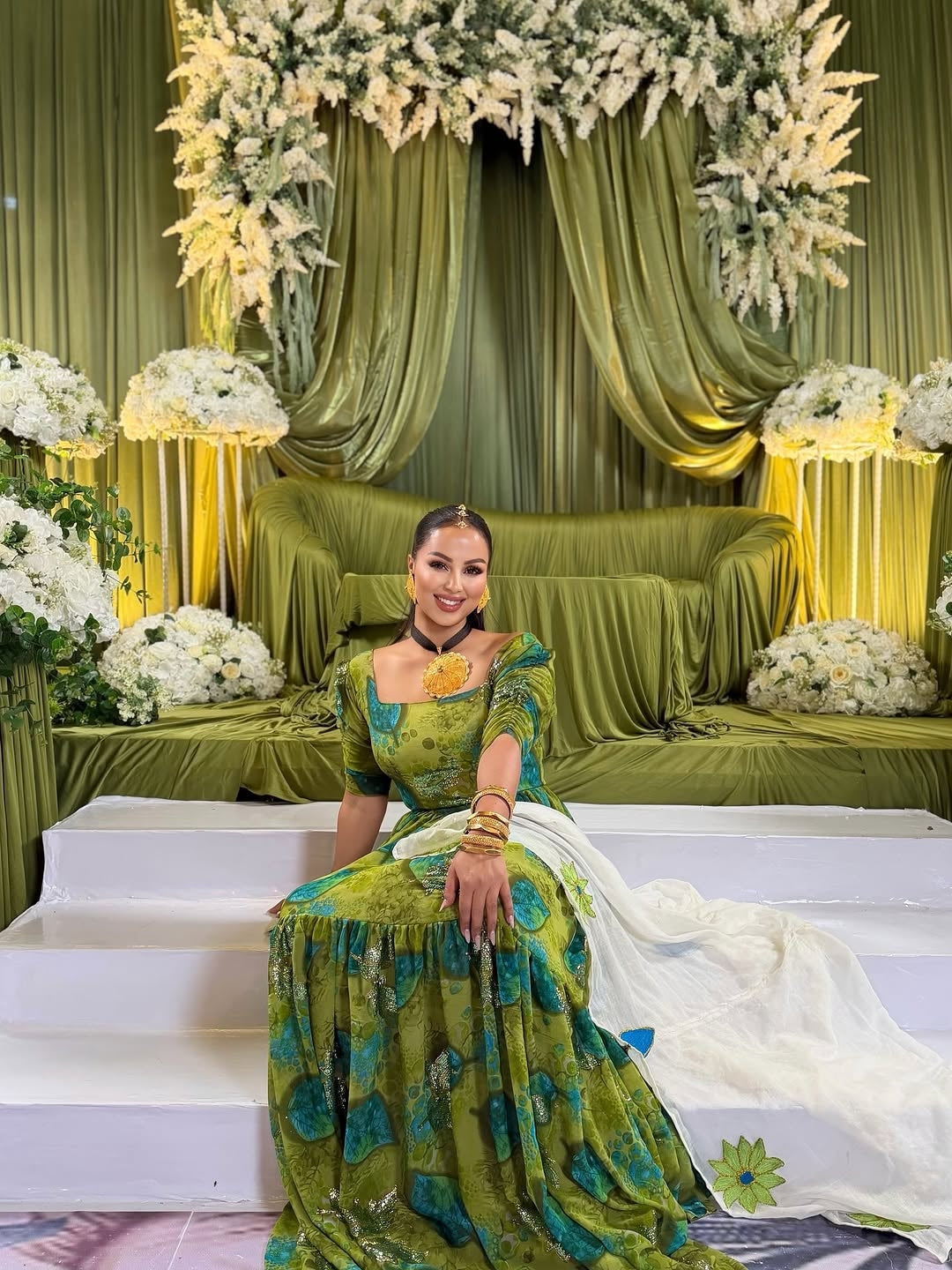 Woman in a green Shiffon Habesha
 sitting on a decorated stage with floral arrangements and green curtains.