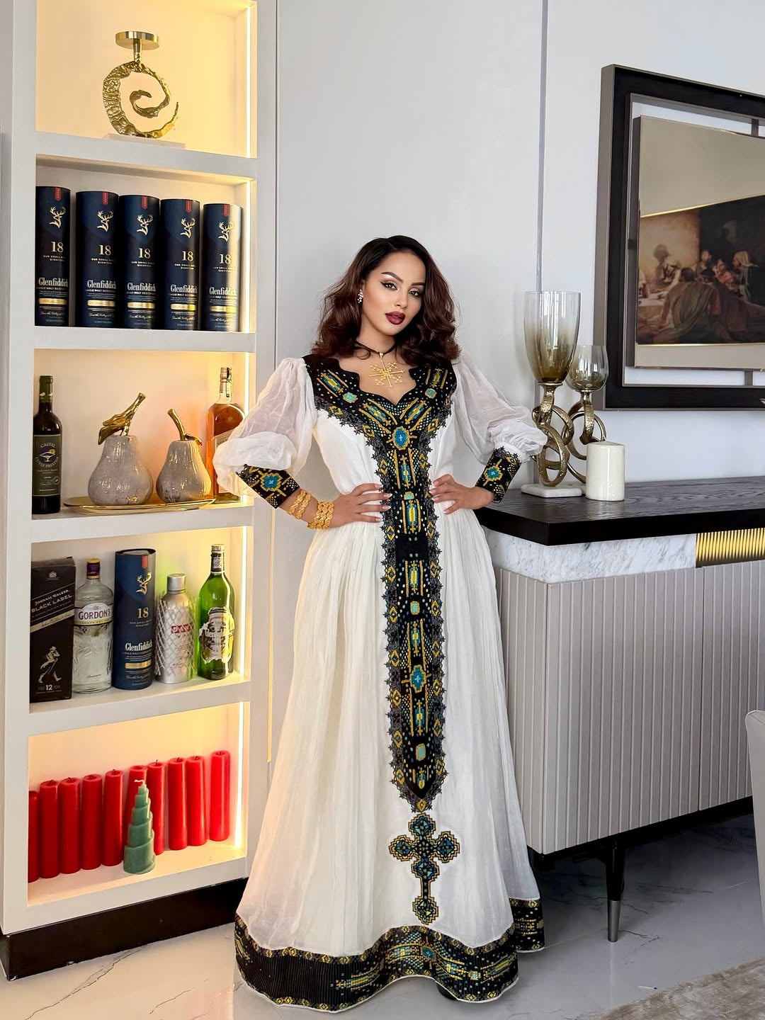 Woman in a white and black embroidered dress standing in a modern interior setting.