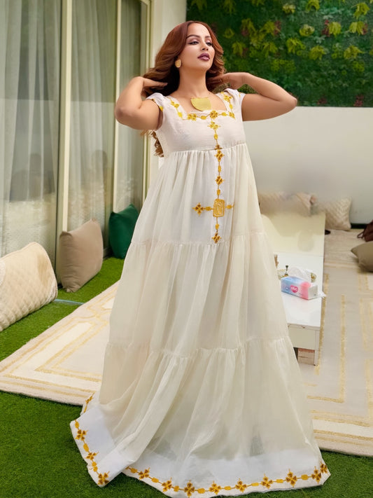 A woman modeling a long white tiered Ethiopian national dresses with yellow floral embroidery, standing in a furnished indoor setting with green artificial grass and cushions.