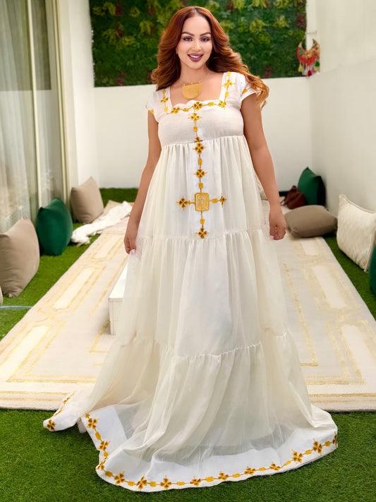 A woman wearing a tiered white maxi Ethiopian national dresses with yellow embroidered details, standing on a green lawn in front of a modern white building.