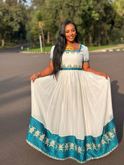 A woman stands outdoors on a paved path wearing a long white habesha kemis with teal blue trim and a decorative border along the hem. She is holding the sides of the skirt outwards to show its full volume.
