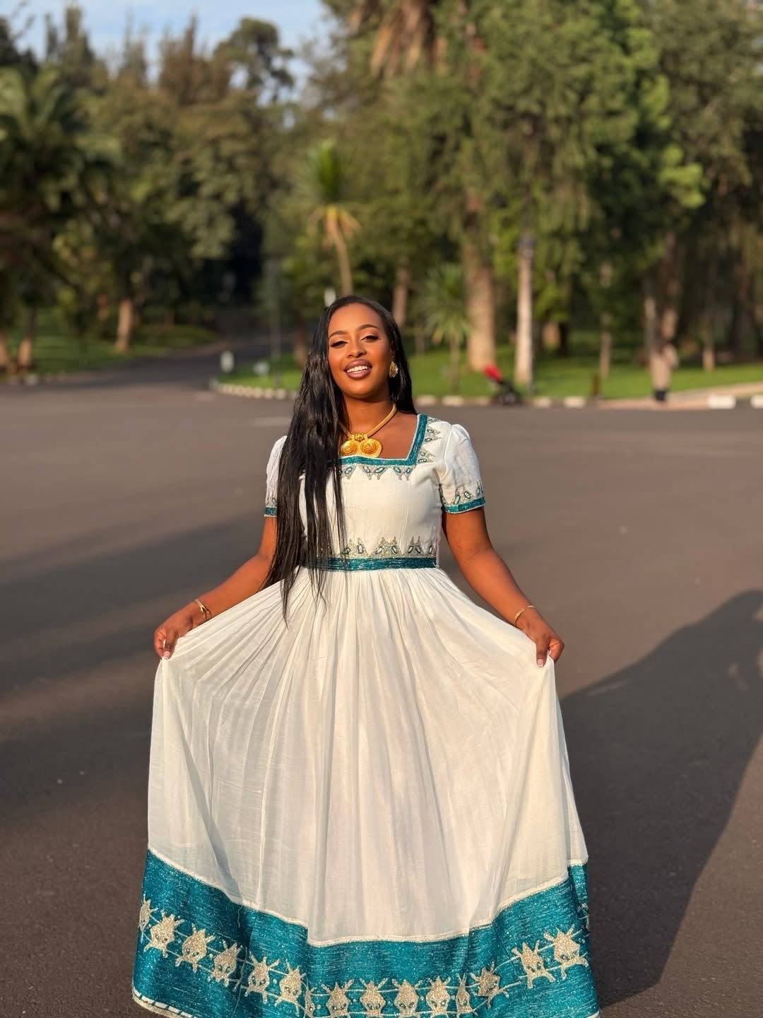 A woman wearing a white and traditional habesha kemis stands outdoors on a paved road, holding out the sides of the skirt to show its full length. The dress features a square neckline, short sleeves with teal trim, a teal waistband, and a wide teal border with white patterns at the hem.