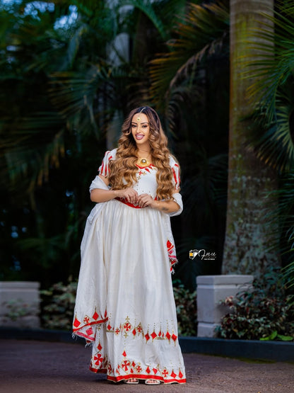 Woman in a white dress with red patterns standing outdoors with greenery in the background