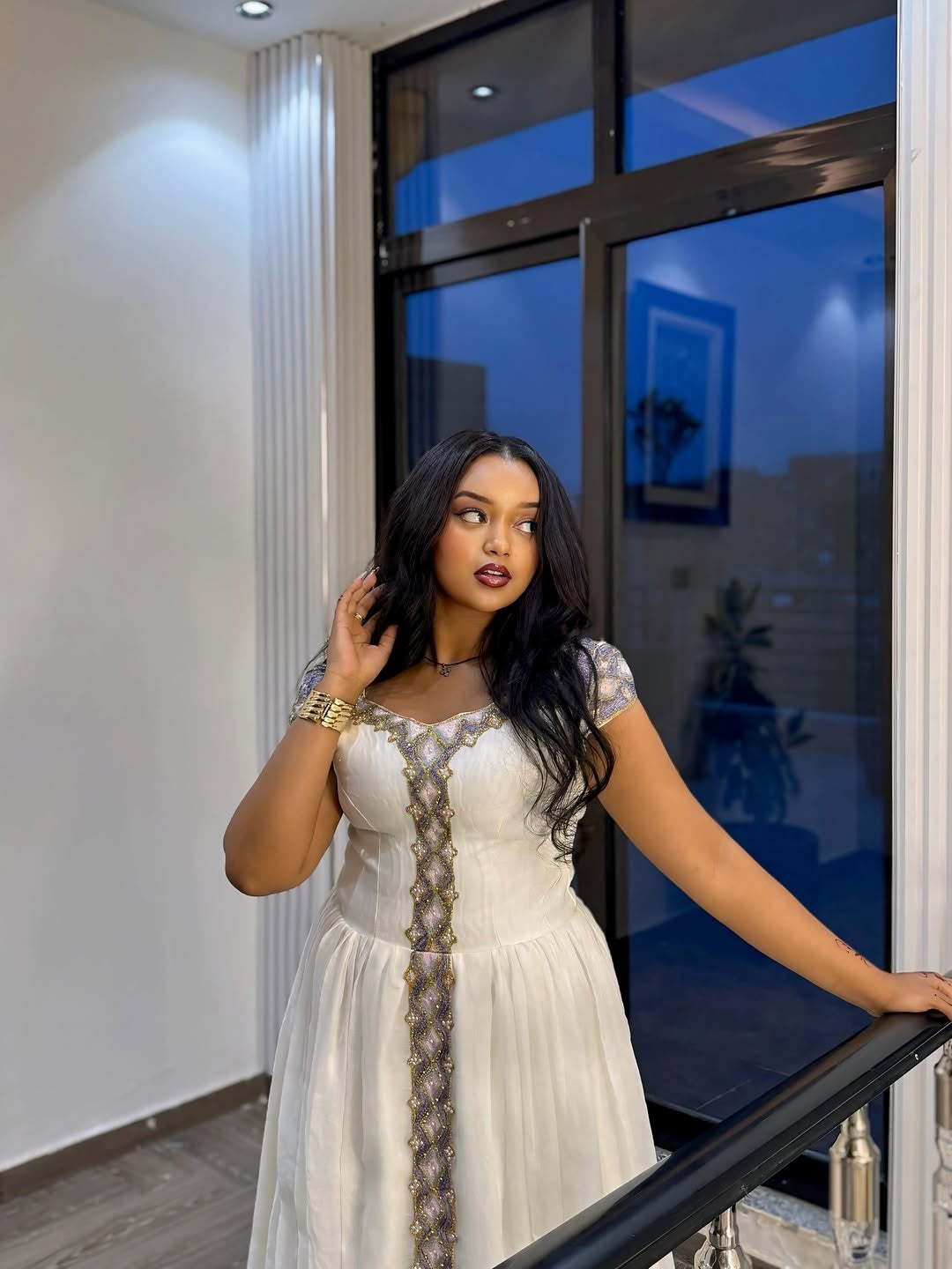 Woman in a white Ethiopian Traditional Dress standing indoors near a large window.