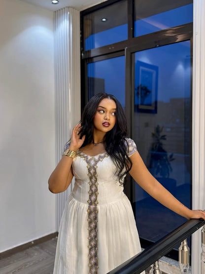 Woman in a white Ethiopian Traditional Dress standing indoors near a large window.