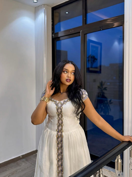 Woman in a white Ethiopian Traditional Dress standing indoors near a large window.