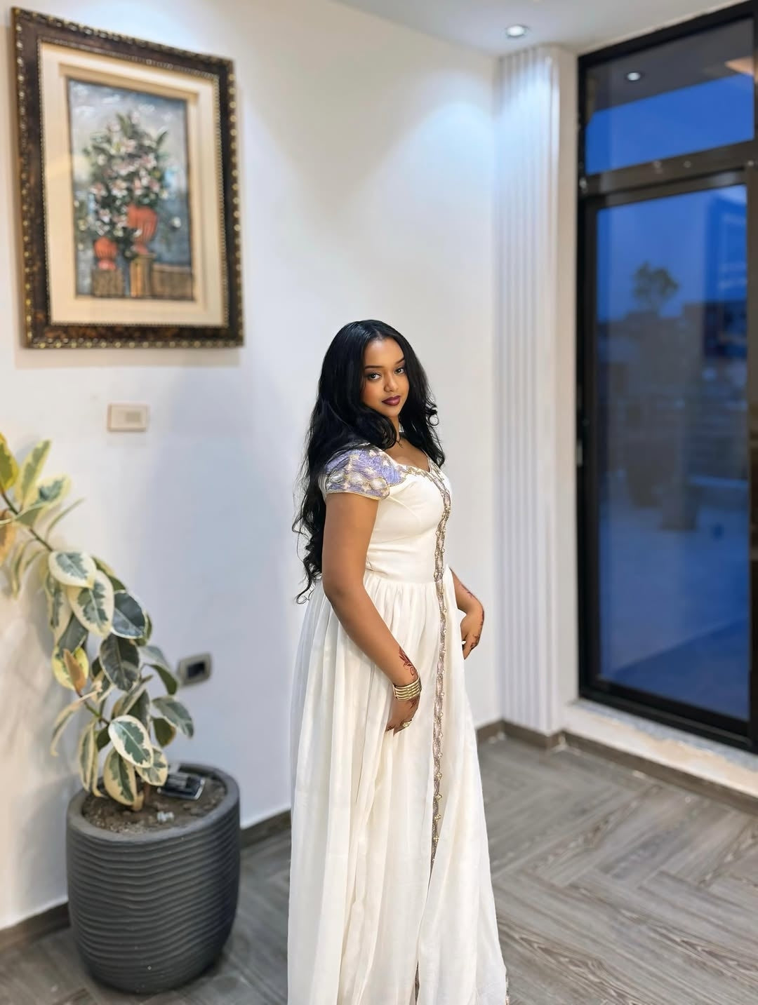 Woman in a white Ethiopian Traditional Dress standing in a room with a painting and plant on the wall.