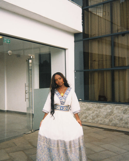  woman standing outdoors in front of a building, wearing a white Habesha dress with patterned blue and yellow embroidery along the V-neckline, waist, and wide hem.