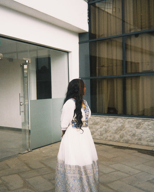 ​"A woman standing outdoors in front of a building, wearing a white Habesha dress with patterned blue and yellow embroidery along the V-neckline, waist, and wide hem."
