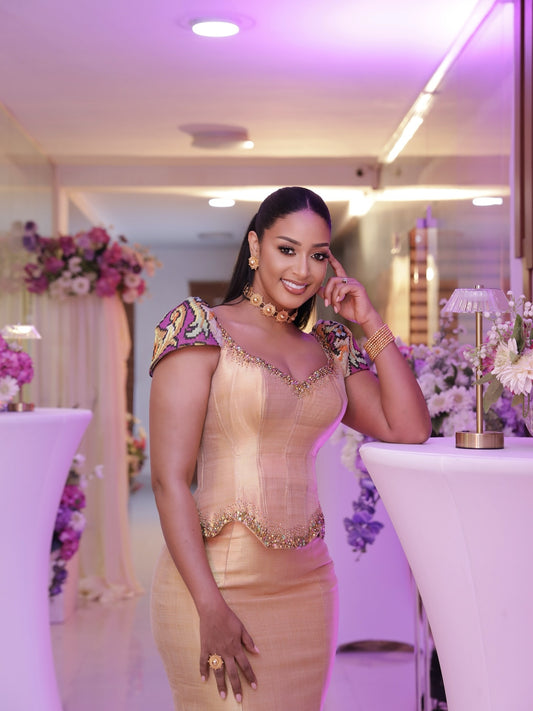 A woman posing in a formal indoor setting wearing a champagne beige traditional-style Traditional Ethiopian Clothing with purple and gold patterned short sleeves and gold jewelry.