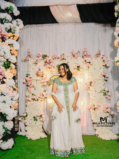 A woman wearing a white traditional habesha kemis with green embroidery stands in front of a decorated backdrop made of white drapery, pink flowers, and artificial green grass. A watermark for 'Mila Pictures' is visible in the lower right corner.