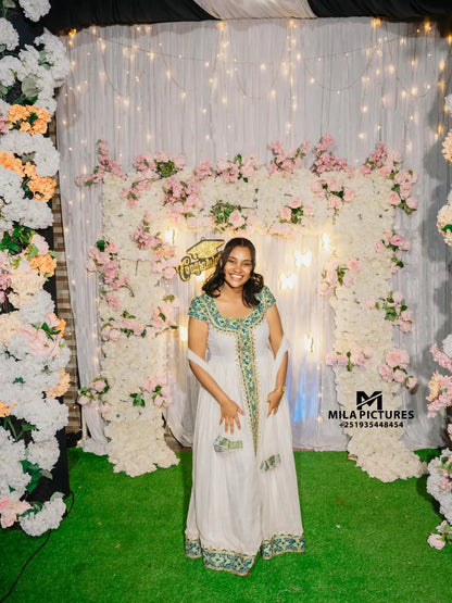 A woman wearing a white traditional habesha kemis with green embroidery stands in front of a decorated backdrop made of white drapery, pink flowers, and artificial green grass. A watermark for 'Mila Pictures' is visible in the lower right corner.