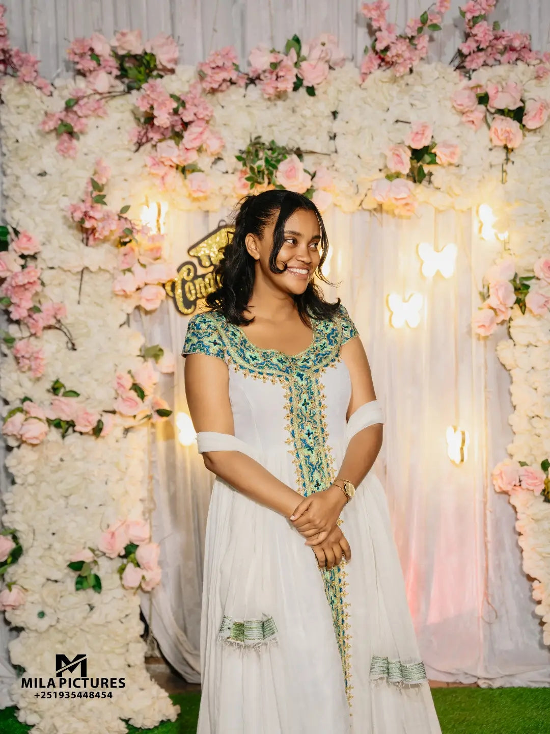A woman wearing a white traditional habesha kemis with green and gold embroidery stands in front of a decorative backdrop made of white flowers, pink roses, and warm string lights. The photo appears to be a wedding or event portrait, with a visible 'Mila Pictures' watermark in the lower left corner.