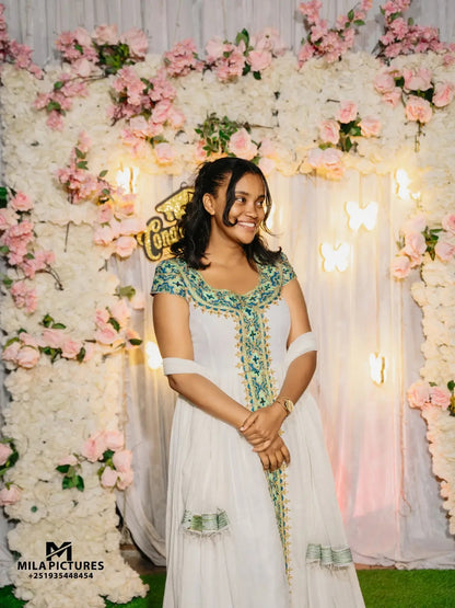 A woman wearing a white traditional habesha kemis with green and gold embroidery stands in front of a decorative backdrop made of white flowers, pink roses, and warm string lights. The photo appears to be a wedding or event portrait, with a visible 'Mila Pictures' watermark in the lower left corner.