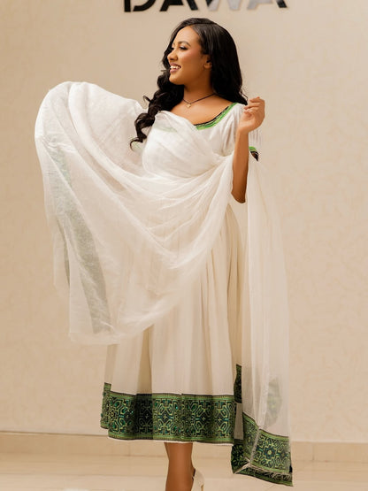 Woman wearing a white Ethiopian Habesha Libs with green border, posing against a beige background
