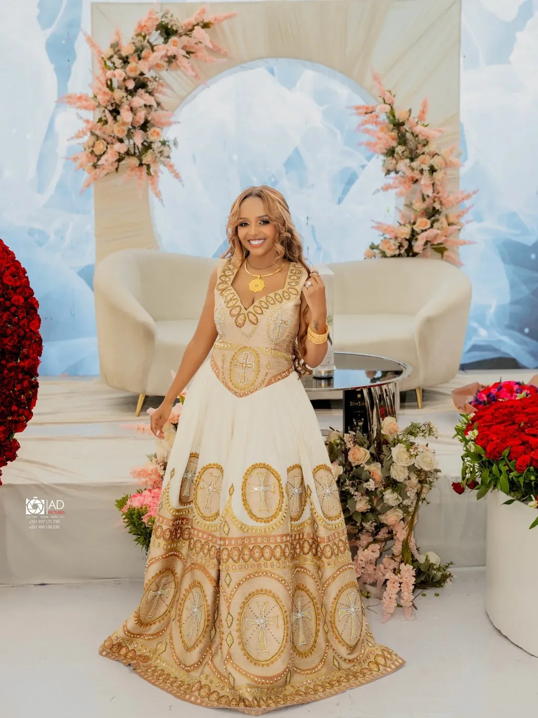 Woman in a gold and white Habesha Libs standing in a decorated room with flowers and a blue abstract wall.
