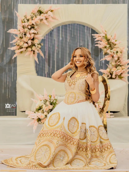 Woman in a gold and white embroidered Habesha Libs posing in front of floral decorations.