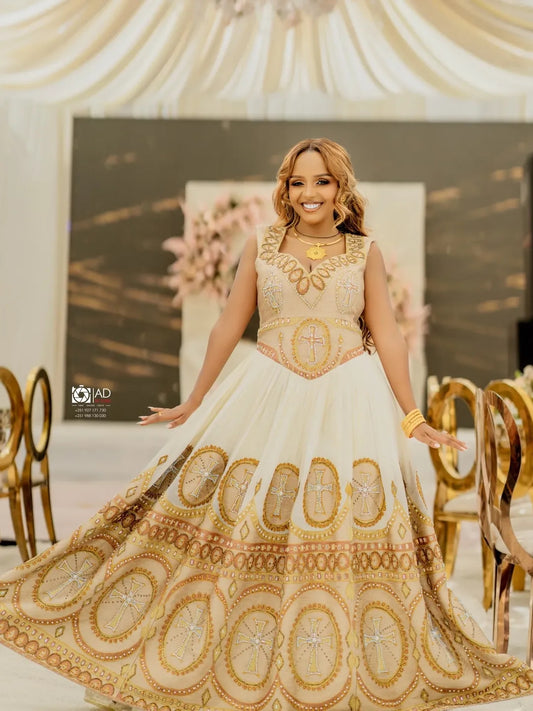 Woman in a white and gold embroidered Habesha Libs standing in an elegant indoor setting.