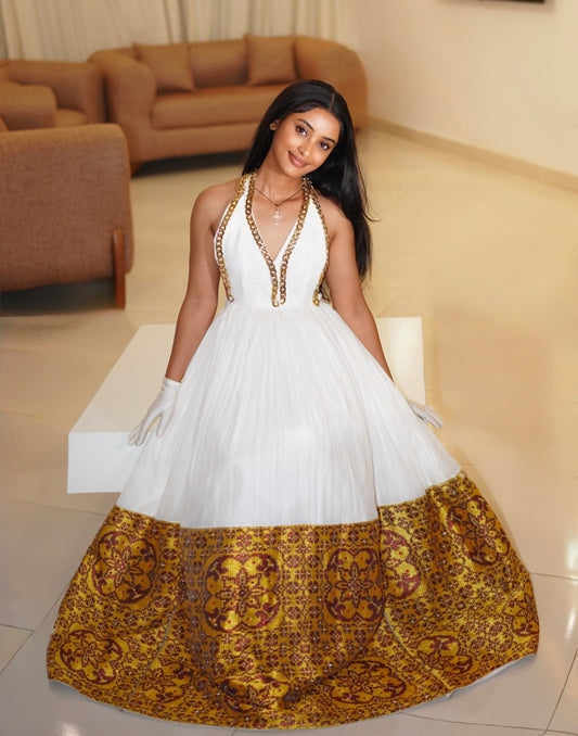A woman wearing a white ball Ethiopian national dresses with a gold patterned hem and gold chain halter details, posing in a modern living room setting.