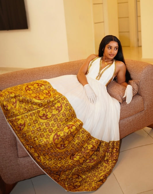 A woman wearing a white sleeveless Ethiopian national dresses with gold trim and white gloves reclines on a brown sofa, with a large yellow and brown patterned fabric draped over the seat and floor in the foreground.