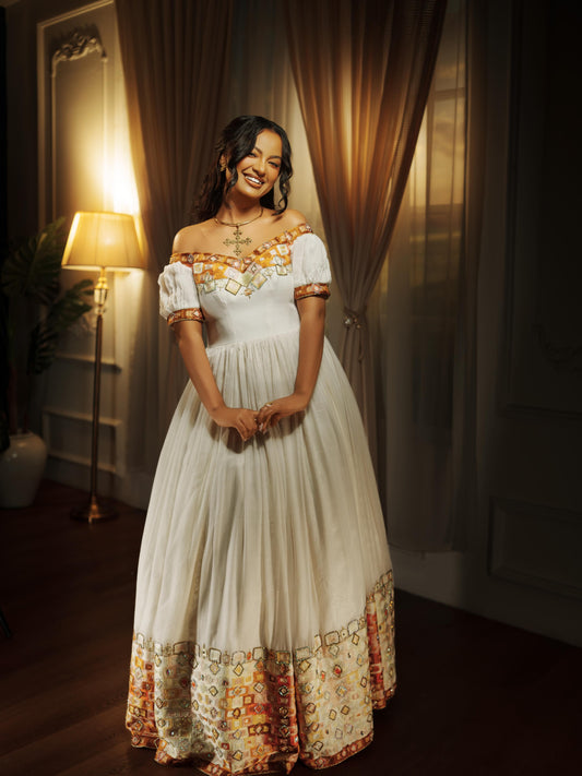 A woman wearing a long off-the-shoulder white Ethiopian national dresses with orange and gold embroidered details and a patterned hem, standing in a warmly lit room with curtains and a floor lamp.