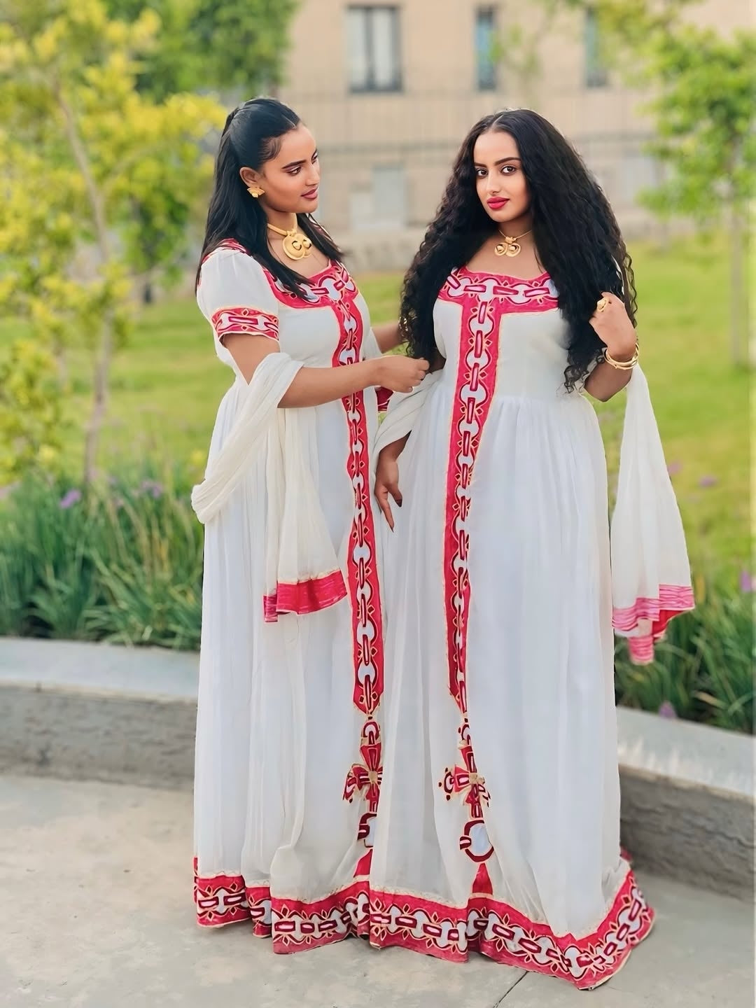 Two women in white and red traditional outfits standing outdoors with greenery in the background.