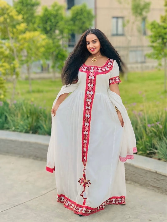 Woman in a white Habesha Zuria with red embroidery standing outdoors.