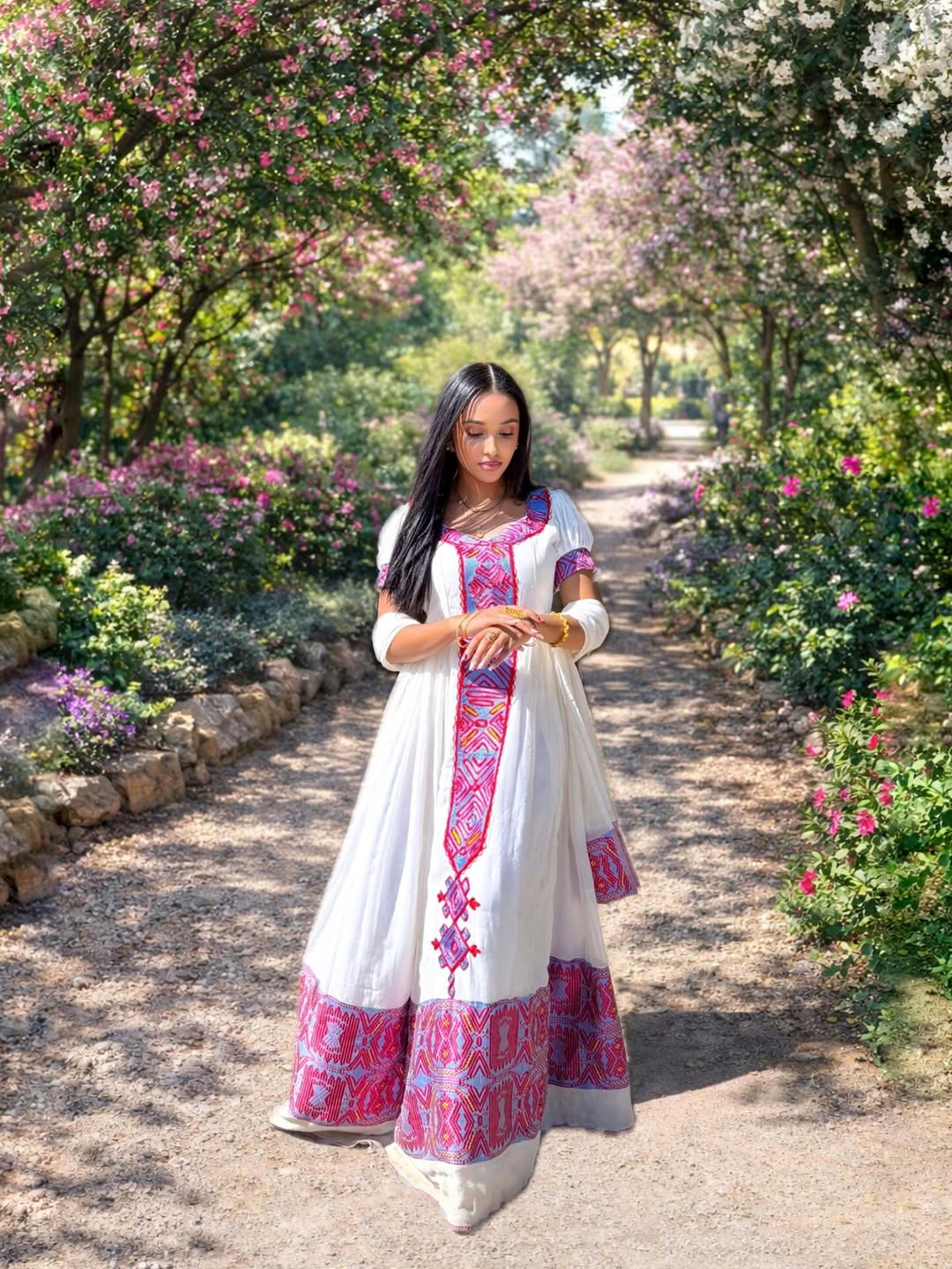 Woman in a habesha dress with pink and purple patterns standing in a garden path.