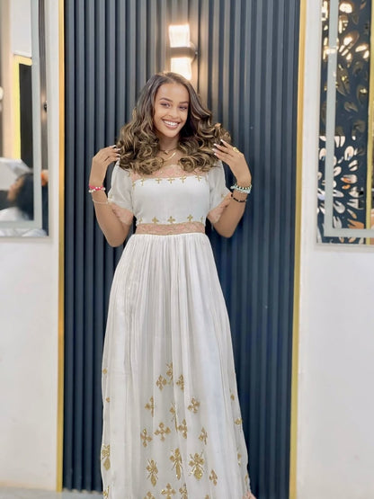 Woman wearing a white dress with gold embroidery in front of a blue curtain.