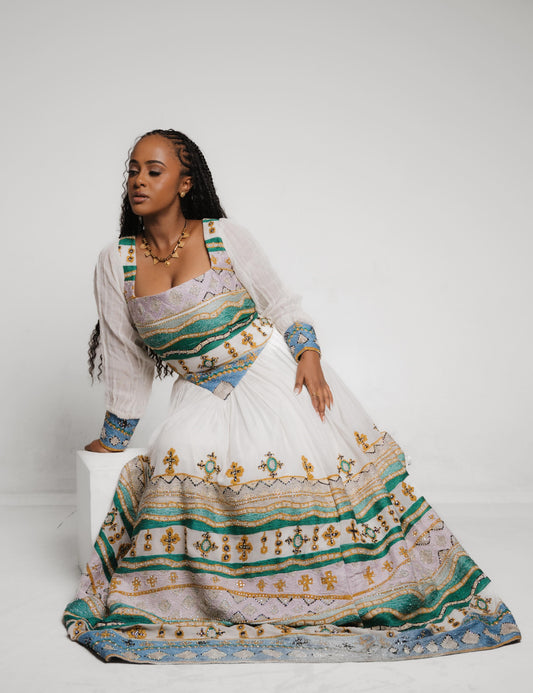 A woman models a traditional long white habesha dress with intricate green, blue, and gold embroidered patterns, posed against a light gray studio background.