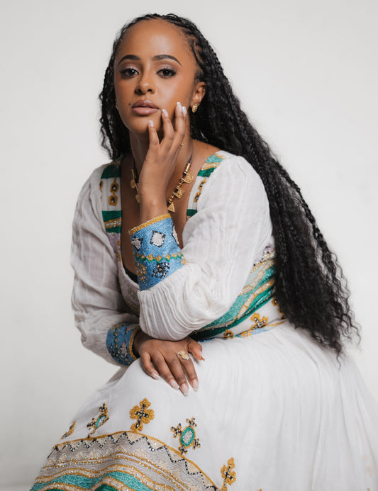 A woman with long black curly hair wearing a white embroidered habesha dress with blue, green, and gold patterns, posing against a plain light gray background.