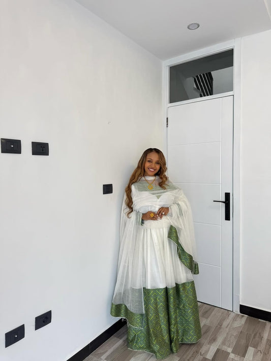 Woman in a white and green Habesha Libs traditional outfit standing in a modern interior setting.
